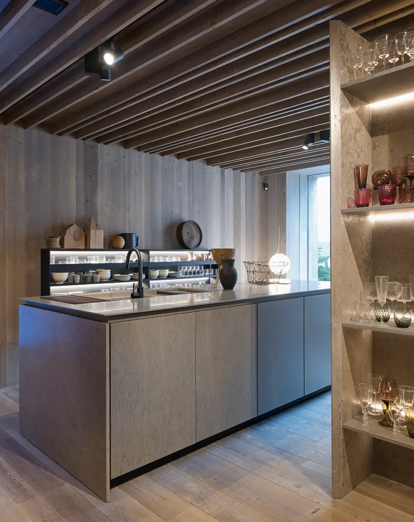 Cucina con isola rivestita in pietra grigia, soffitto a travi di legno e mensole illuminate con bicchieri e oggetti in vetro.