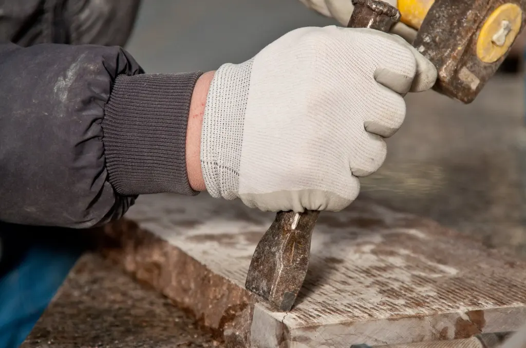 Mano con guanto da lavoro che lavora il bordo di una lastra di pietra con scalpello e mazzetta.
